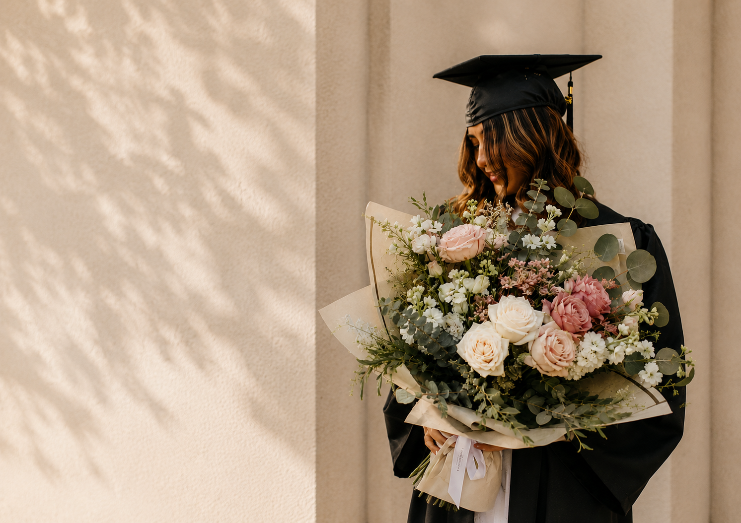 Young woman in black graduation cap and gown holding a large bouquet of pink, white, and cream roses with eucalyptus